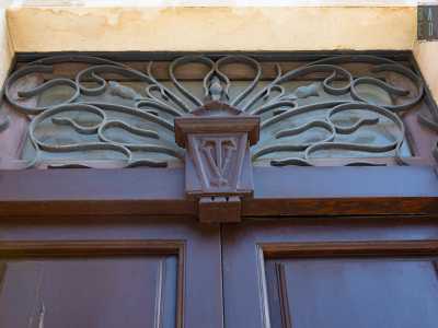 Sullo scuro portone in legno capeggiano le iniziali del costruttore scolpite in un decoro ligneo, sovrastato da una griglia geometrica